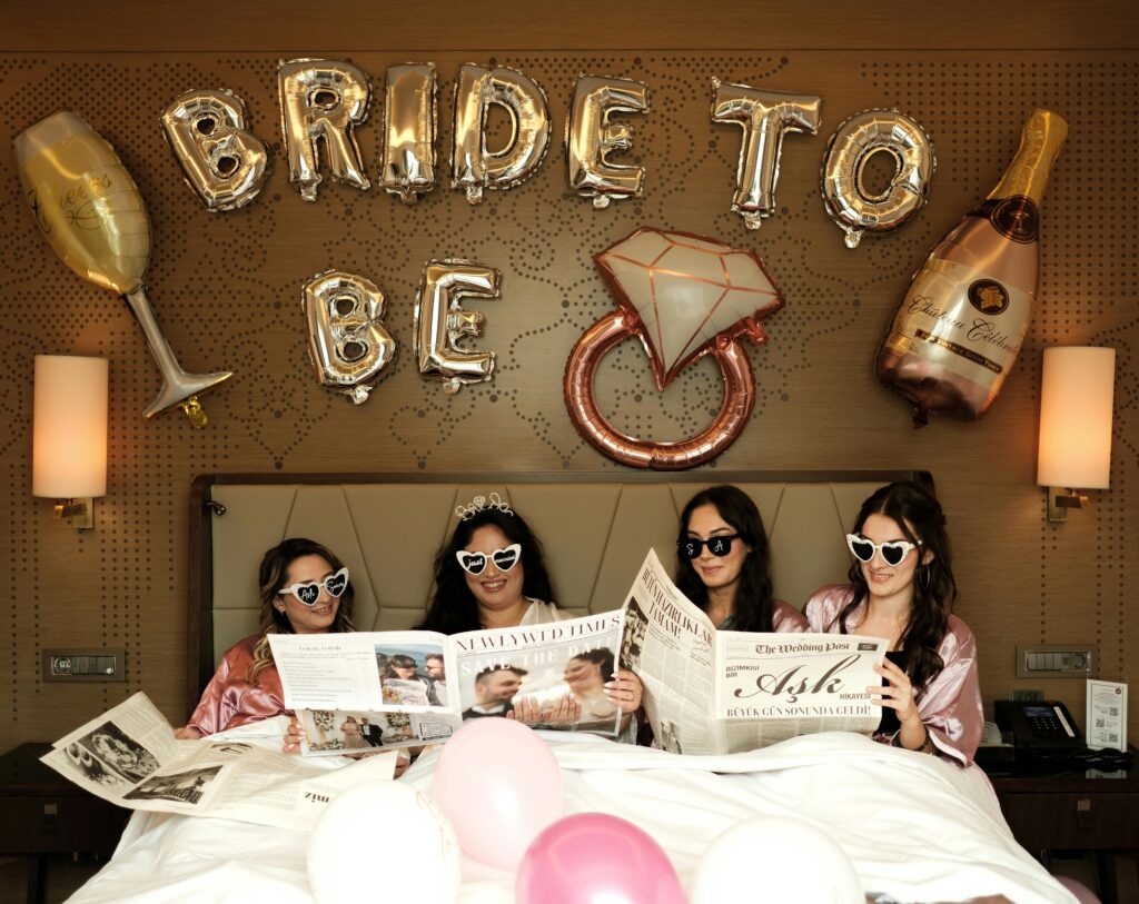 Four women celebrating a bachelorette party in a hotel room with fun props and balloons.