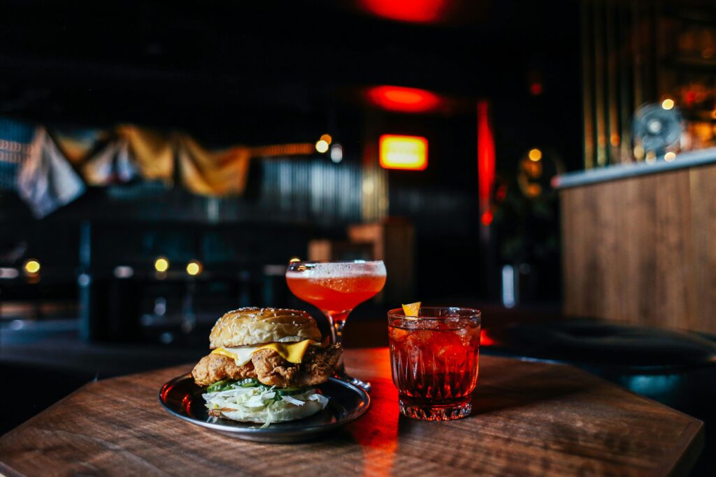 Close-up of a gourmet burger with cocktails on a table in a dimly lit bar, creating a cozy atmosphere.